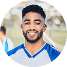 Smiling young man with beard in a white and blue sports jersey outdoors.