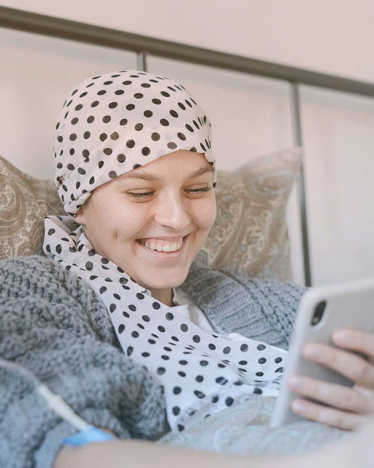 Junge Krebspatientin mit Kopftuch lächelt im Bett und schaut aufs Smartphone – Symbol für Lebensfreude und Zuversicht während der Krebstherapie mit unterstützender Behandlung wie CBD oder THC.