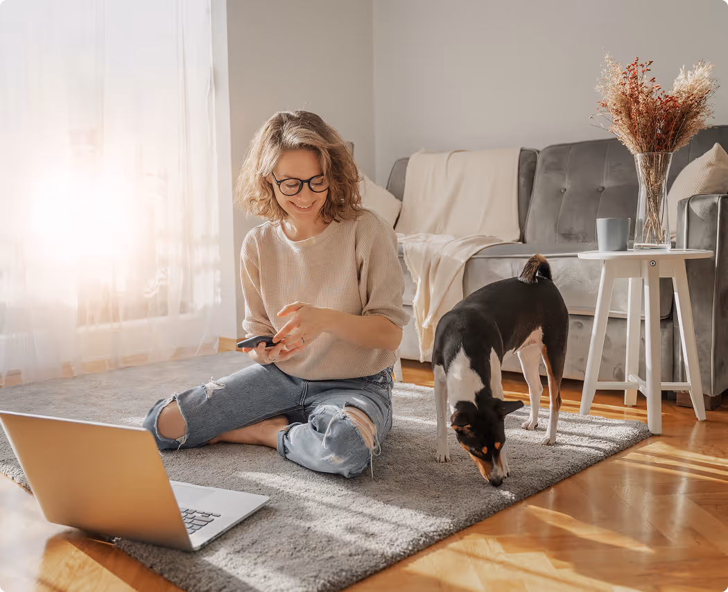 Woman wearing glasses sitting cross-legged on a gray rug using a smartphone with a laptop in front and a black and white dog sniffing the floor nearby.