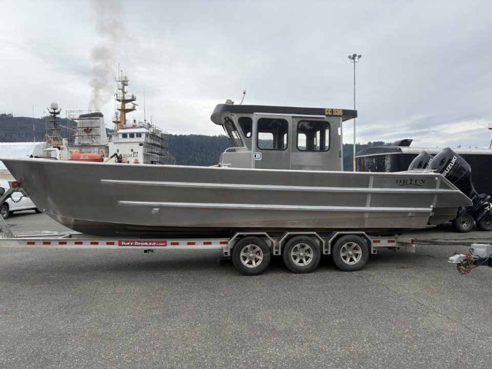 Ortun aluminum cabin cruiser boat on a triple-axle trailer at the dock, cargo ship in background.