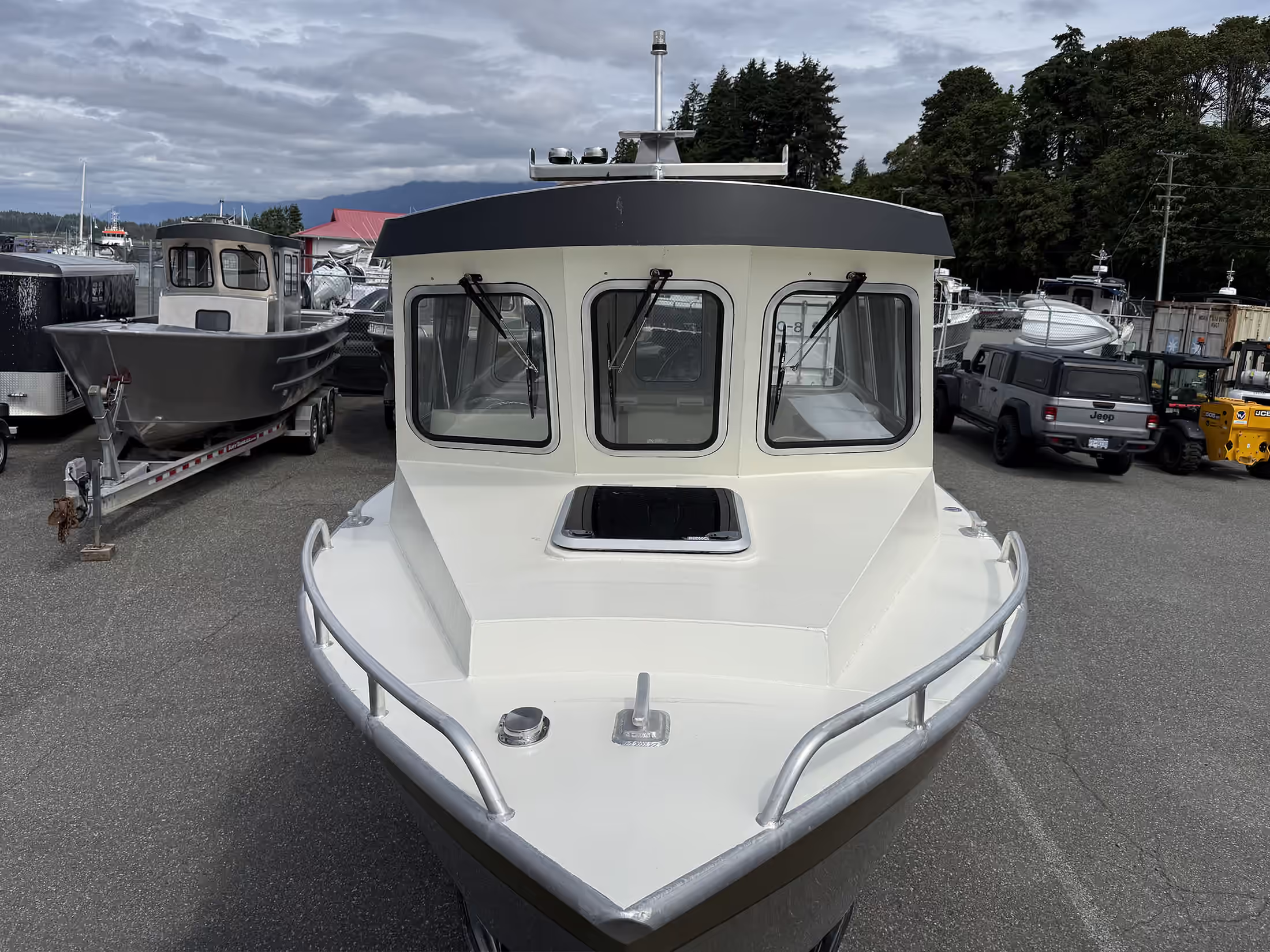 Front view of a white and black cabin boat with three windshield windows, metal railings on the deck, and a cloudy sky in the background, parked in a lot with other boats and vehicles.