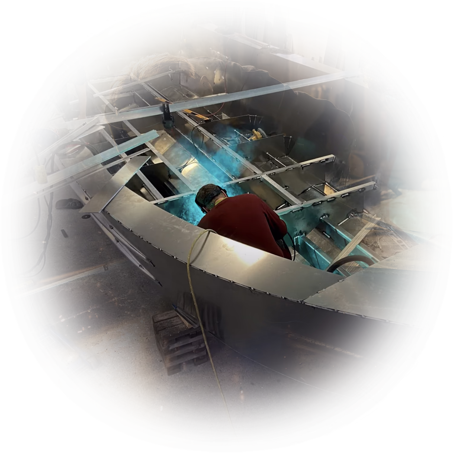 A person welding metal inside a partially constructed boat frame in a workshop, with blue welding light illuminating the area.