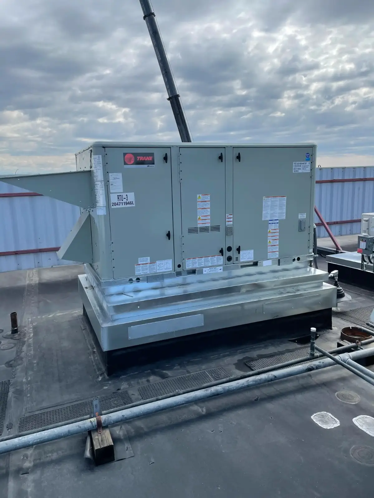 A large, light-grey Trane rooftop unit (RTU-3) with external ductwork sits on a metal curb on a flat roof under a cloudy sky.