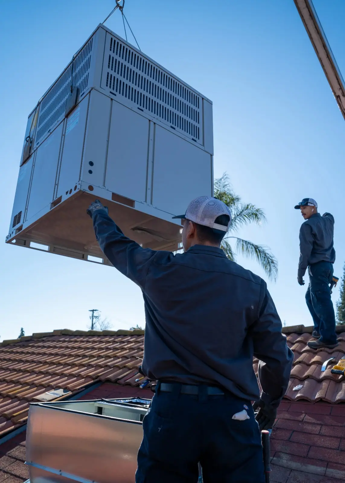 Two technicians oversee the aerial lowering of a large HVAC unit onto a pitched residential roof, with a crane visible above and blue sky in the background.