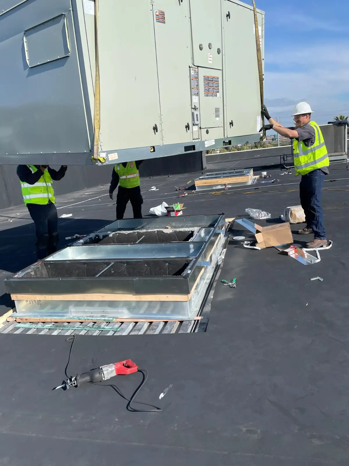 Three construction workers in safety vests and hard hats guide a large, white rooftop HVAC unit being lifted by a crane onto a metal curb on a black roof.