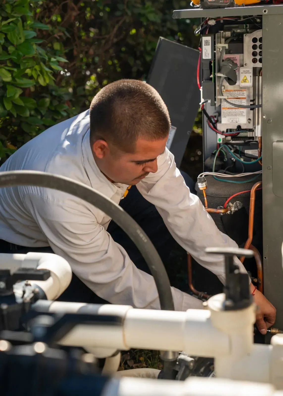 Technician kneeling outdoors, carefully working with wires inside an open HVAC unit surrounded by greenery