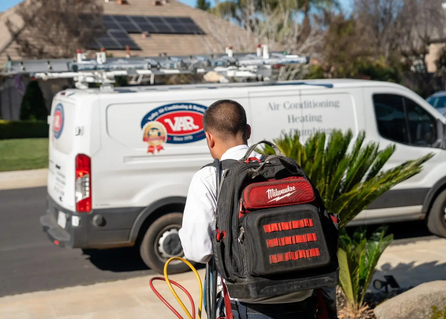 HVAC technician carrying a tool backpack and refrigerant hoses walking toward a Valley Air service van.