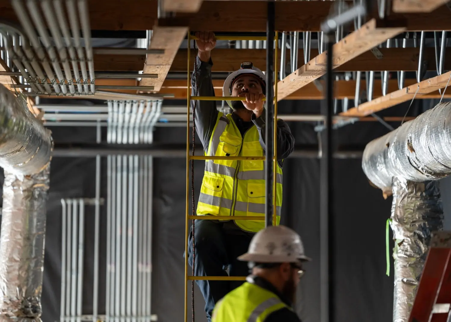 Construction worker in hard hat and safety vest climbing ladder to inspect overhead pipes and electrical conduit.