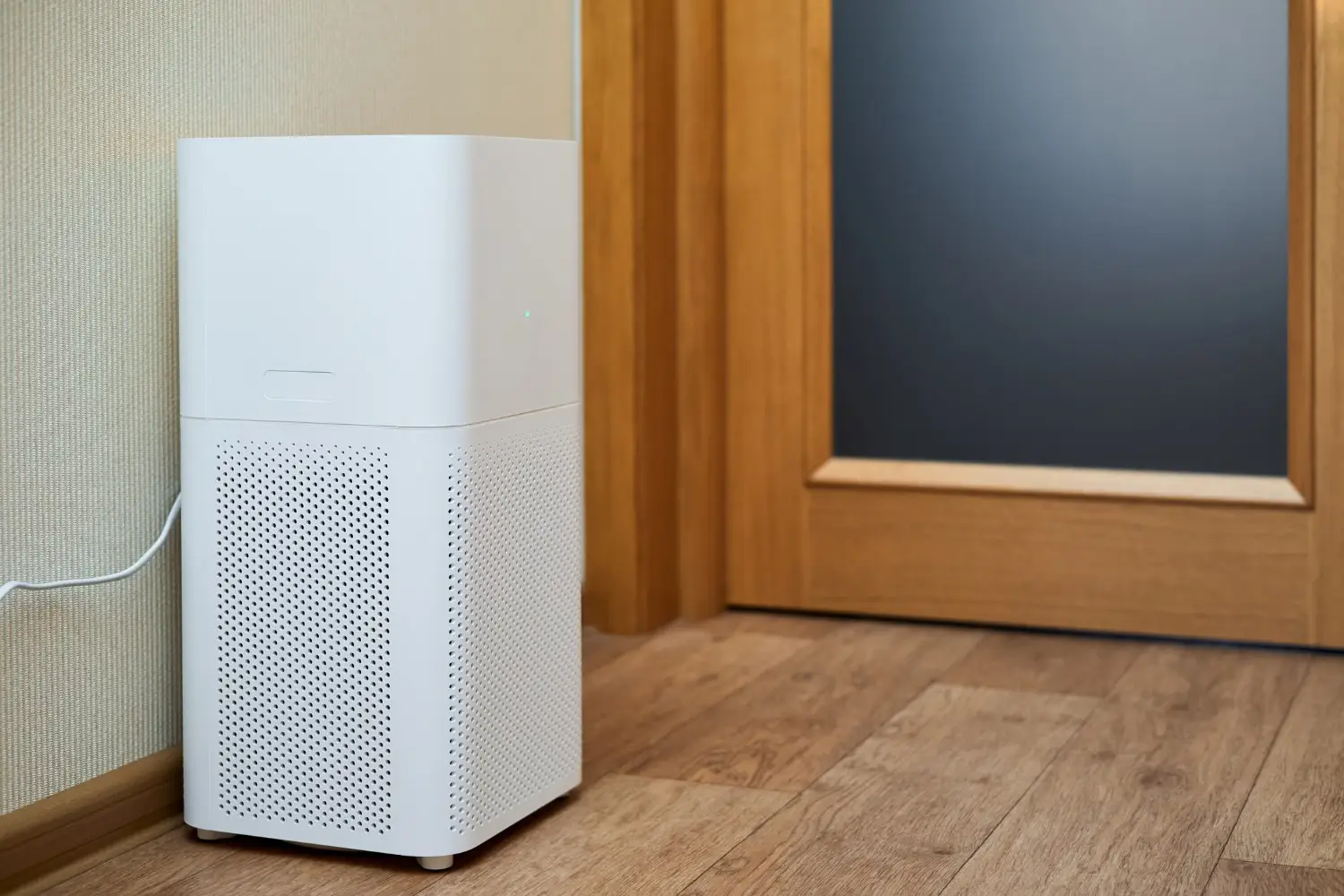 A tall, white air purifier sits on a wooden floor near a wall and a wooden door with a dark glass panel.