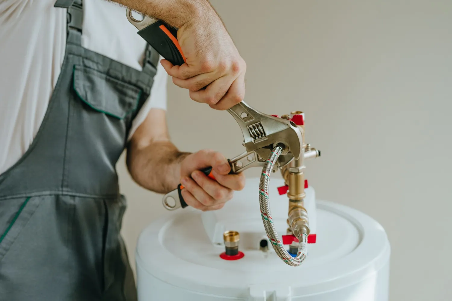 A technician in grey overalls is using a large adjustable wrench to tighten a flexible braided hose connection on top of a white water heater or boiler.
