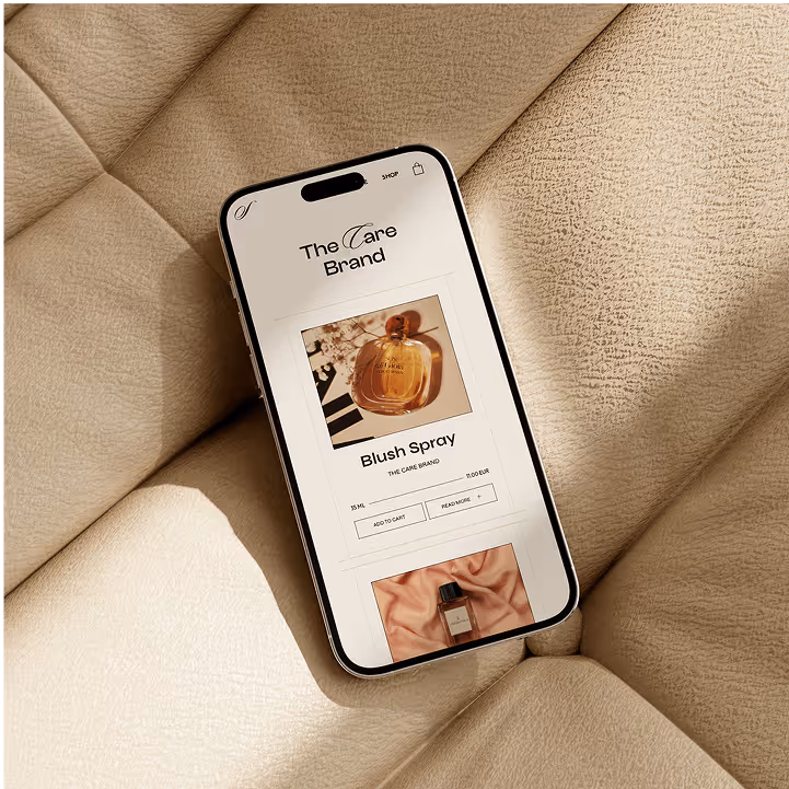 Smartphone zeigt eine Website von The Care Brand mit einem Bild eines Blush Spray Parfüms auf einem beigen, gepolsterten Hintergrund.
