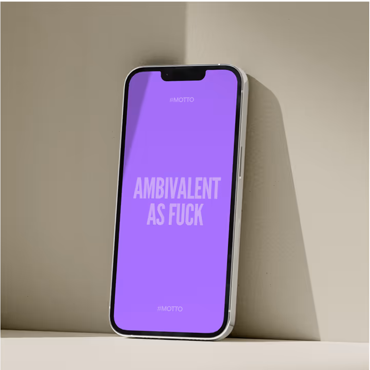 Ein Smartphone lehnt an einer cremefarbenen Wand, der Bildschirm zeigt auf lila Hintergrund den Text ‚Ambivalent as fuck‘.