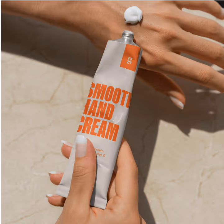Hand hält eine Tube mit oranger Aufschrift 'Smooth Hand Cream' und drückt weiße Handcreme auf die andere Hand.