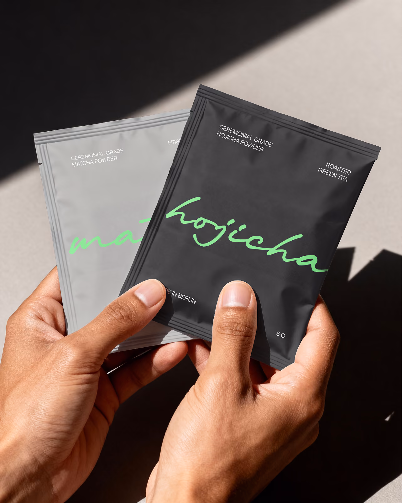 Person hält zwei kleine Beutel: ein grauer Matcha-Pulver- und ein schwarzer Hojicha-Pulverbeutel.