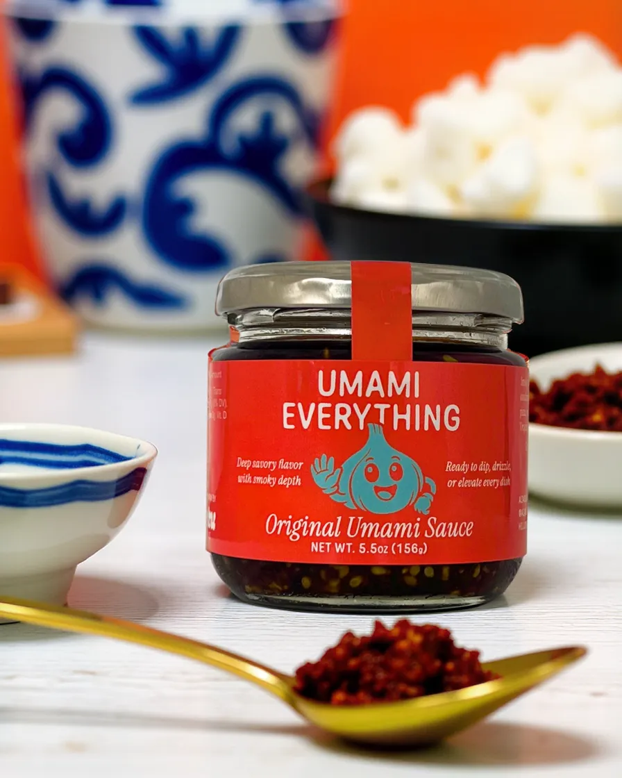 Jar of Azabu Foods’ Original Umami Everything Sauce placed on a white surface with a gold spoonful of sauce in front, surrounded by blue-and-white ceramic bowls and a bowl of rice in the background, highlighting the product’s vibrant packaging and everyday kitchen versatility.
