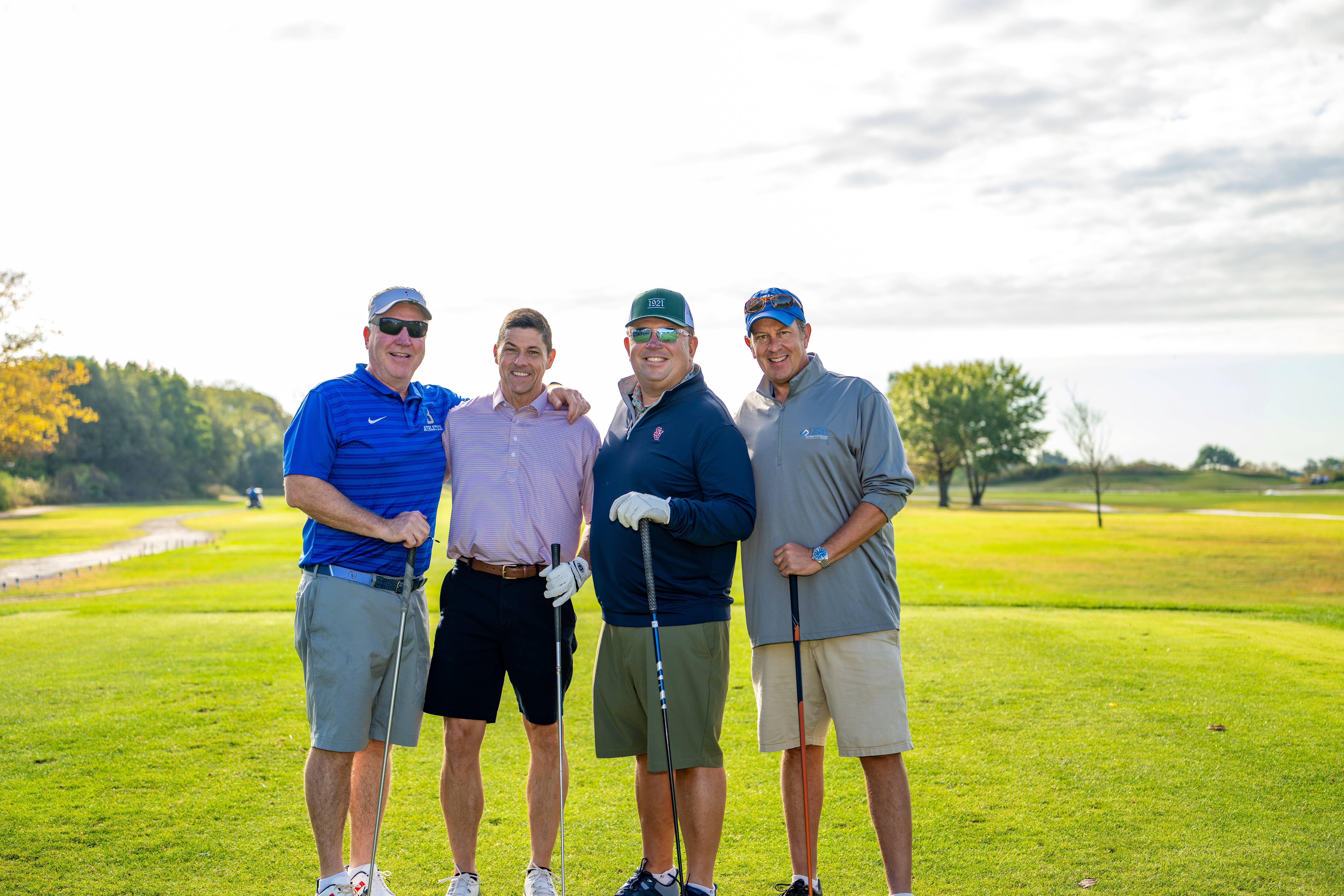 Four males posing with golf clubs on golf course.