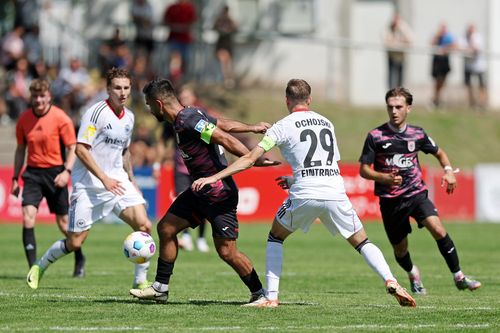 Fußballspiel mit zwei Spielern in schwarzen Trikots und einem Spieler in weißem Trikot mit der Aufschrift Ochojski 29, die um den Ball kämpfen.