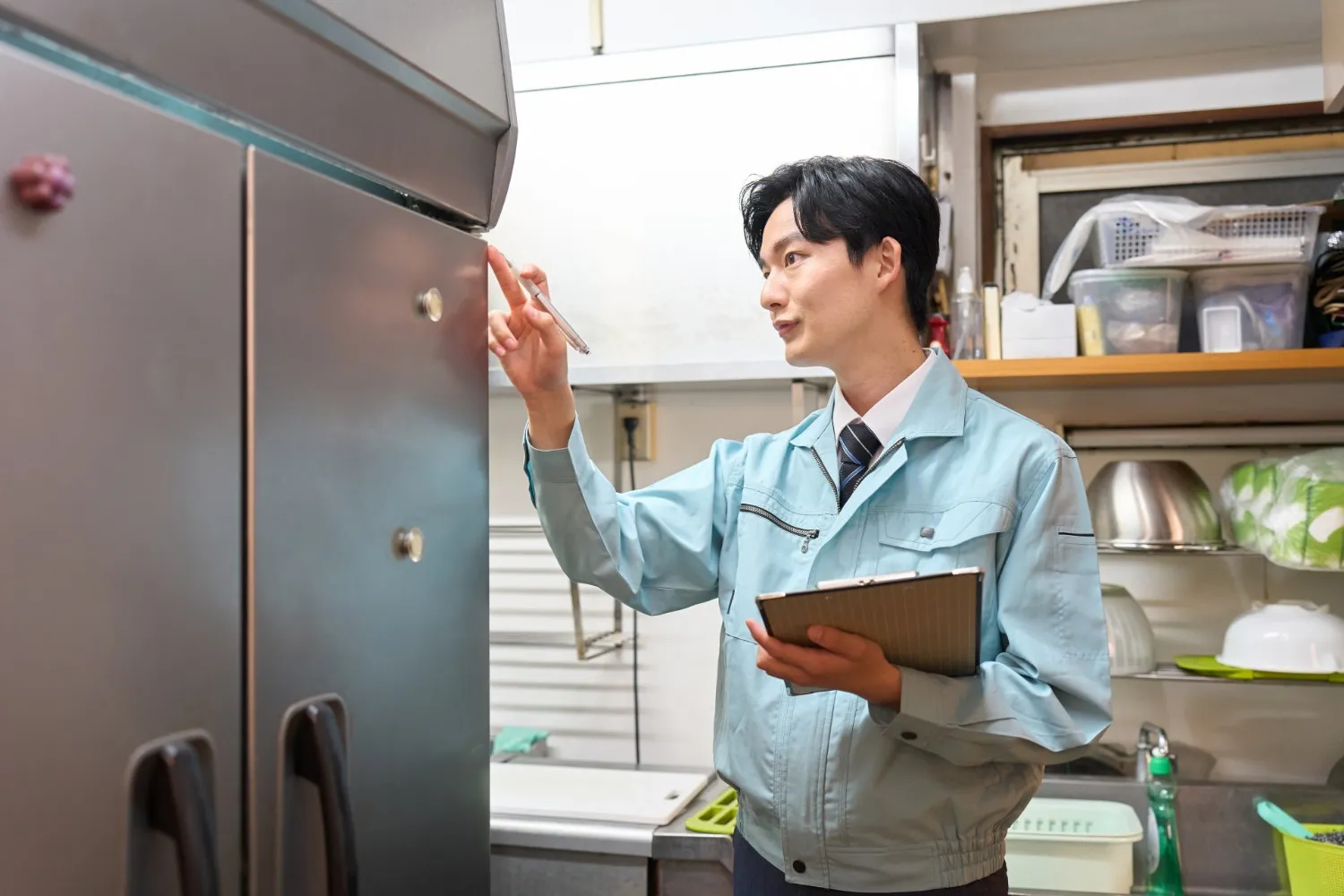 A young male technician in a light blue work uniform and tie holds a clipboard and inspects a large, industrial-style stainless steel refrigerator or freezer. He uses a pen to point at the unit's exterior in what appears to be a commercial kitchen or storage area.