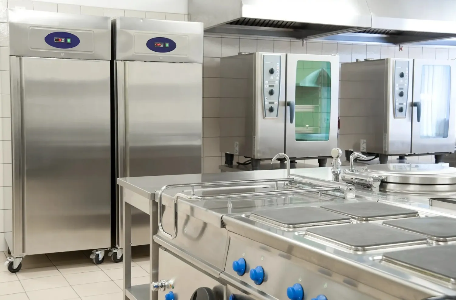 Commercial kitchen with stainless steel refrigerators, combination ovens, and a cooking range with a work surface.