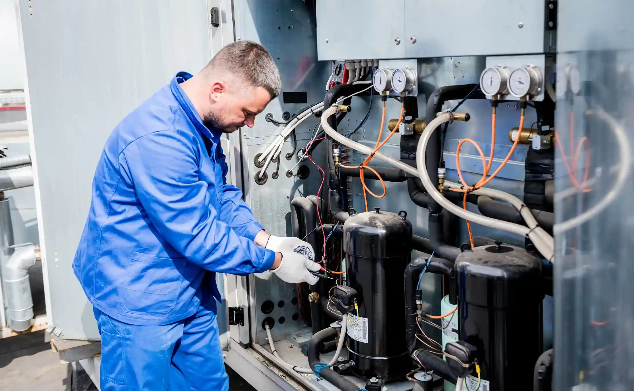 A professional technician in a blue uniform and white gloves inspects the compressor and coils of an industrial refrigeration unit.