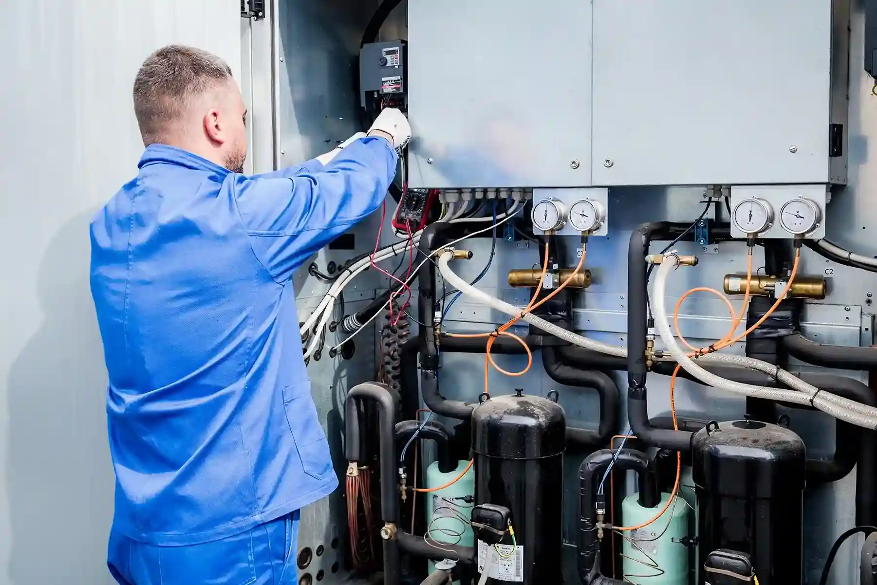 A service technician in a blue uniform and gloves works on the complex pipes and components of a commercial HVAC or refrigeration system.