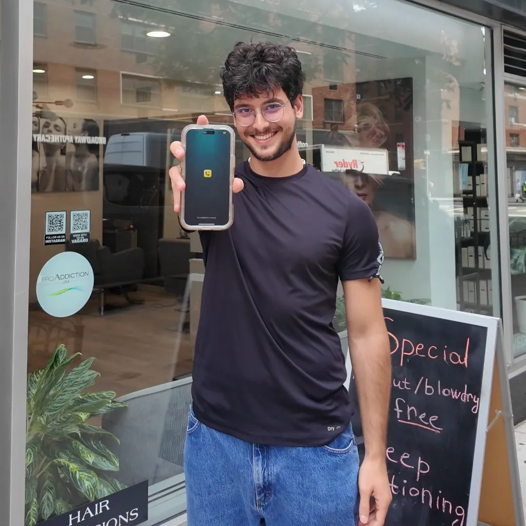 Smiling man with glasses holding a smartphone showing an app screen, standing outside a hair salon.