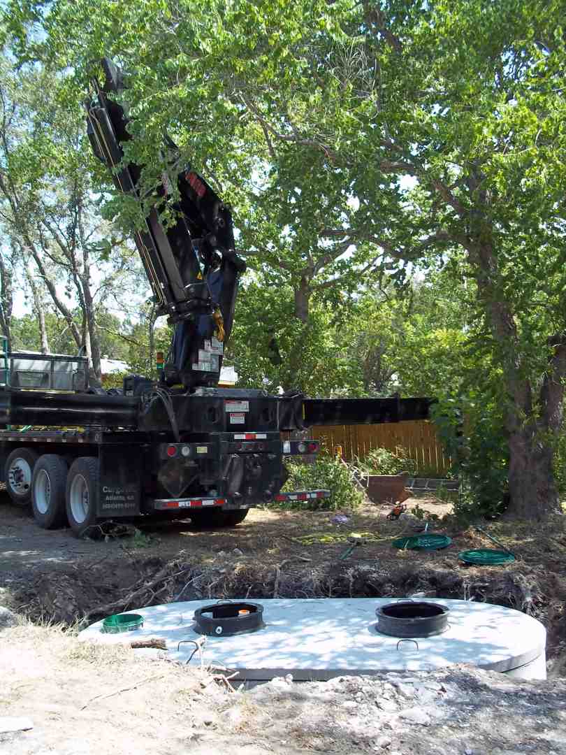 A septic tank being lowered into the ground by an excavator, with workers in safety gear and hard hats, a red and white safety vest is visible - install aerobic septic unit san antonio tx