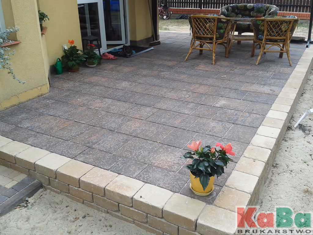 Brukowany taras z donicą pełną czerwonych kwiatów i meblami ogrodowymi na końcu.