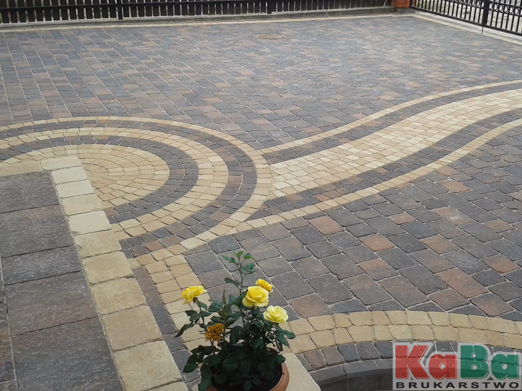 Podwórko z brukowaną nawierzchnią w odcieniach beżu i szarości z donicą żółtych róż na pierwszym planie.