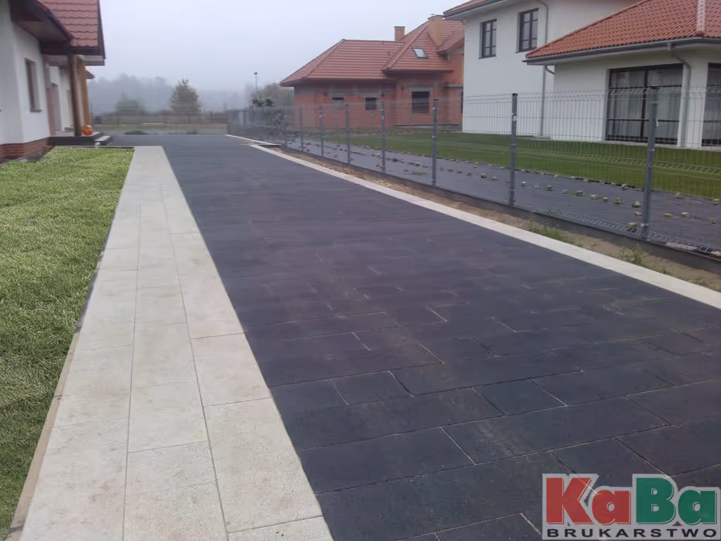 Podjazd wyłożony ciemnymi, prostokątnymi płytkami z białą ścieżką i zielonym trawnikiem obok, z domami w tle.