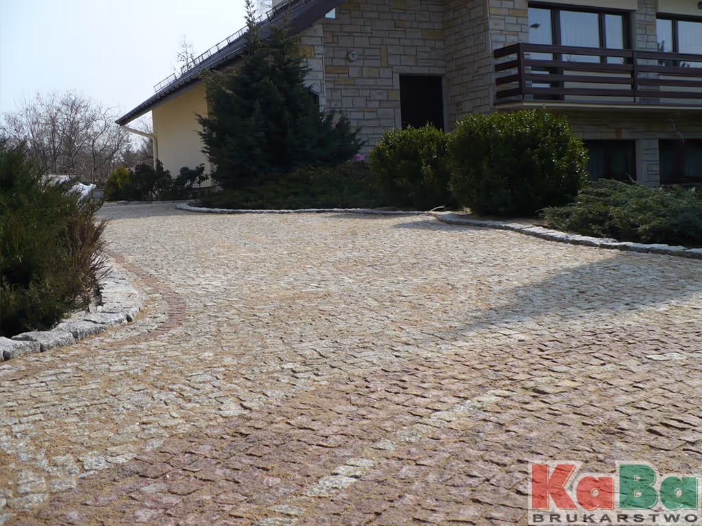 Podjazd do domu wyłożony nieregularną kostką granitową o różnych odcieniach szarości i różu, otoczony zielonymi krzewami i drzewami.