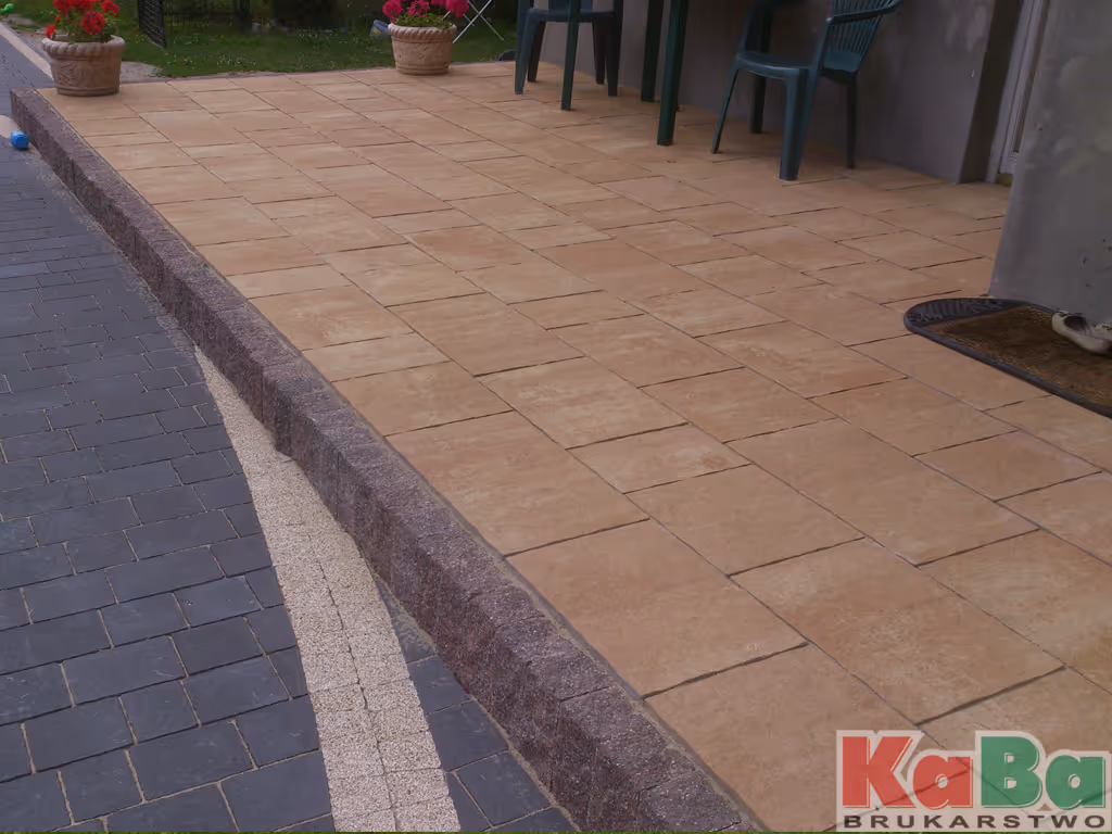 Taras wyłożony beżowymi kwadratowymi płytkami z podwyższeniem oddzielającym go od ciemnej kostki brukowej i donicami z czerwonymi kwiatami.