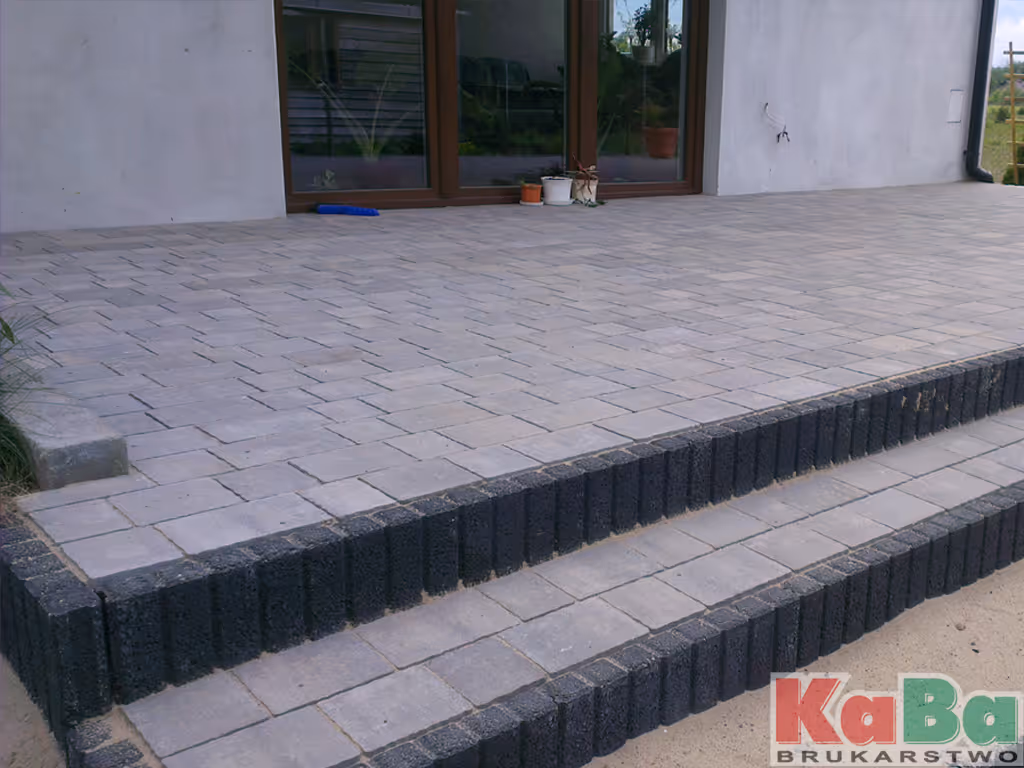 Taras z jasnoszarych płytek brukowych z ceglanym obramowaniem o grafitowym kolorze oraz schodami prowadzącymi do patio.