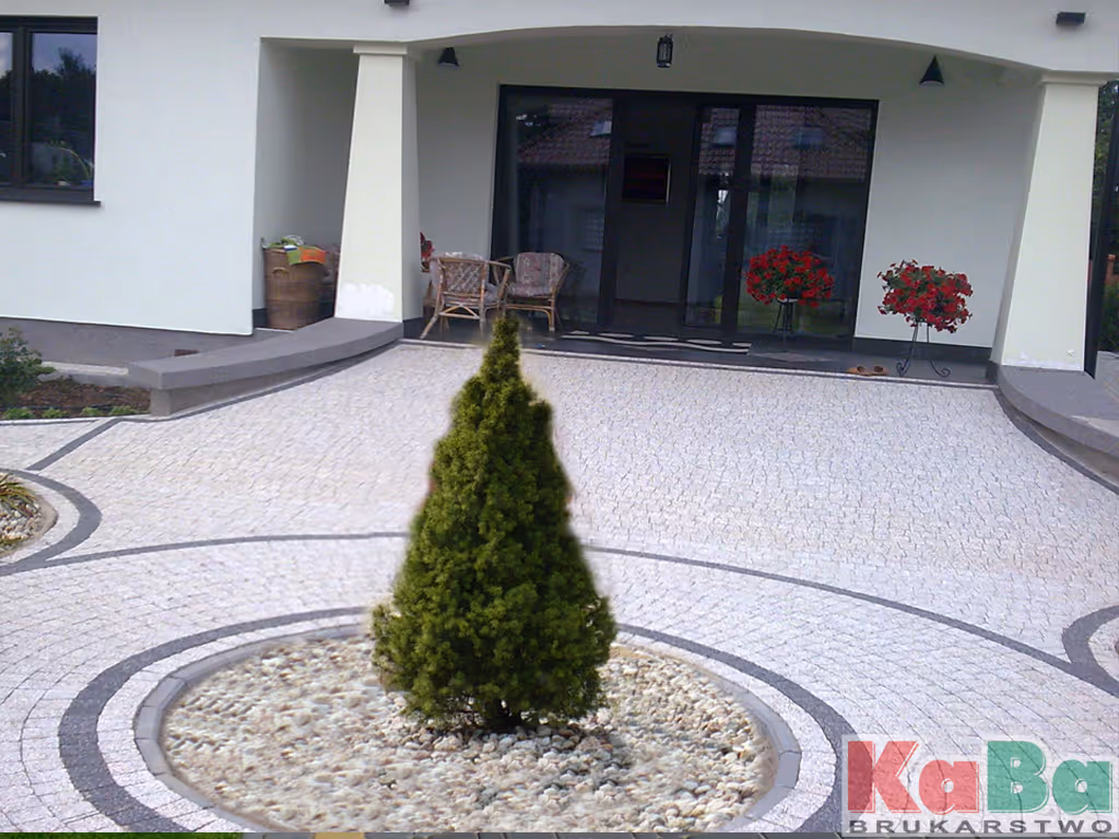 Podjazd wyłożony jasnymi kostkami z centralnie umieszczonym zielonym krzewem na tle wejścia do białego domu z dwoma fotelami i czerwonymi kwiatami.