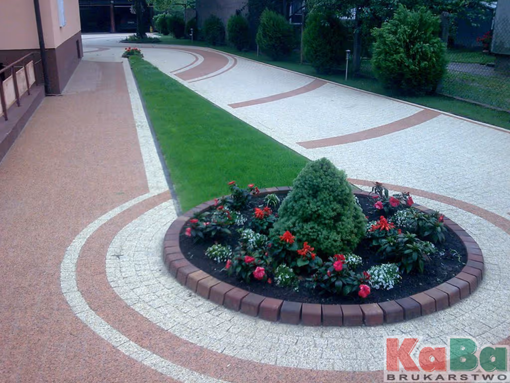 Ozdobny okrągły klomb z małym drzewkiem i czerwonymi kwiatami na tle brukowanej ścieżki i zielonego trawnika.