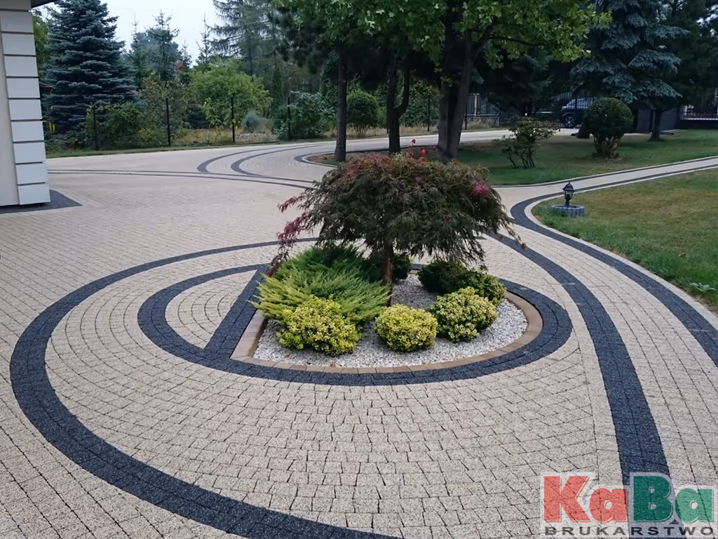 Ogród z kamiennym chodnikiem z geometrycznym wzorem i małym drzewem otoczonym zielonymi krzewami.