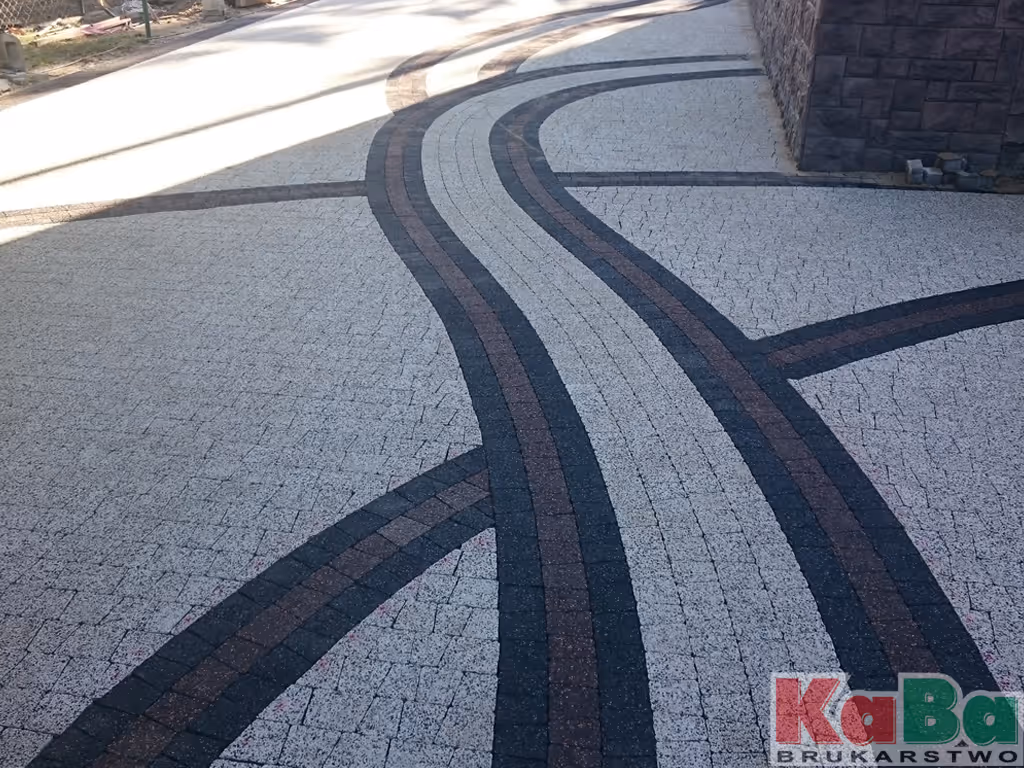 Układ brukowanych kamieni w jasnych i ciemnych odcieniach tworzący wzór zakręcającej ścieżki przy budynku.