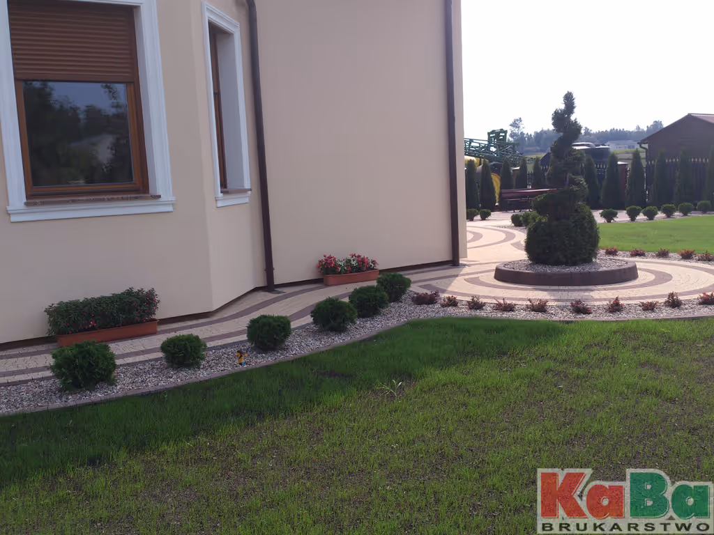 Ogród z zielonym trawnikiem, krzewami i podjazdem z dekoracyjnym wzorem przy jasnym budynku.