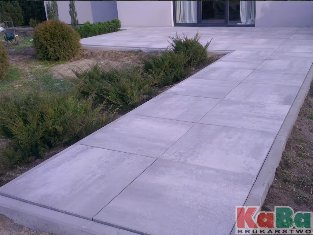 Popielaty chodnik i podest wykonane z dużych prostokątnych płyt betonowych obok zielonego ogrodu przed wejściem do domu.