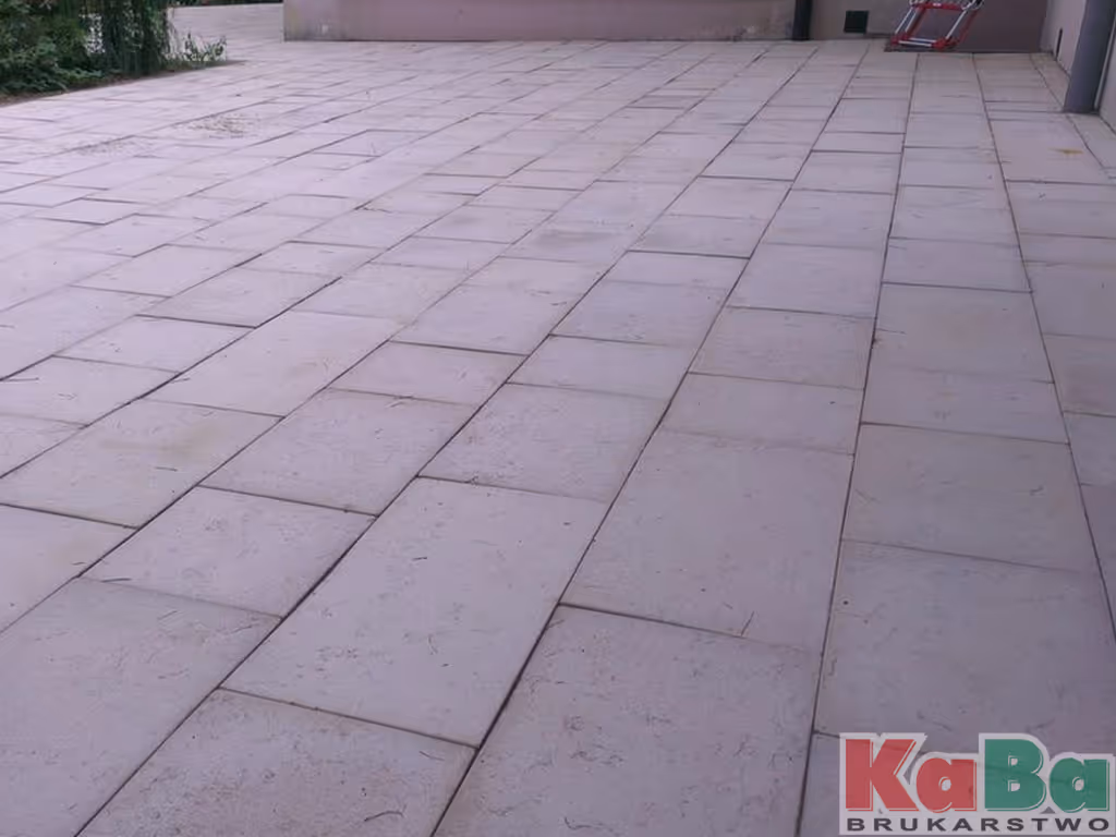 Duży taras wyłożony jasnoszarymi, prostokątnymi płytkami betonowymi ułożonymi w przesunięte rzędy.