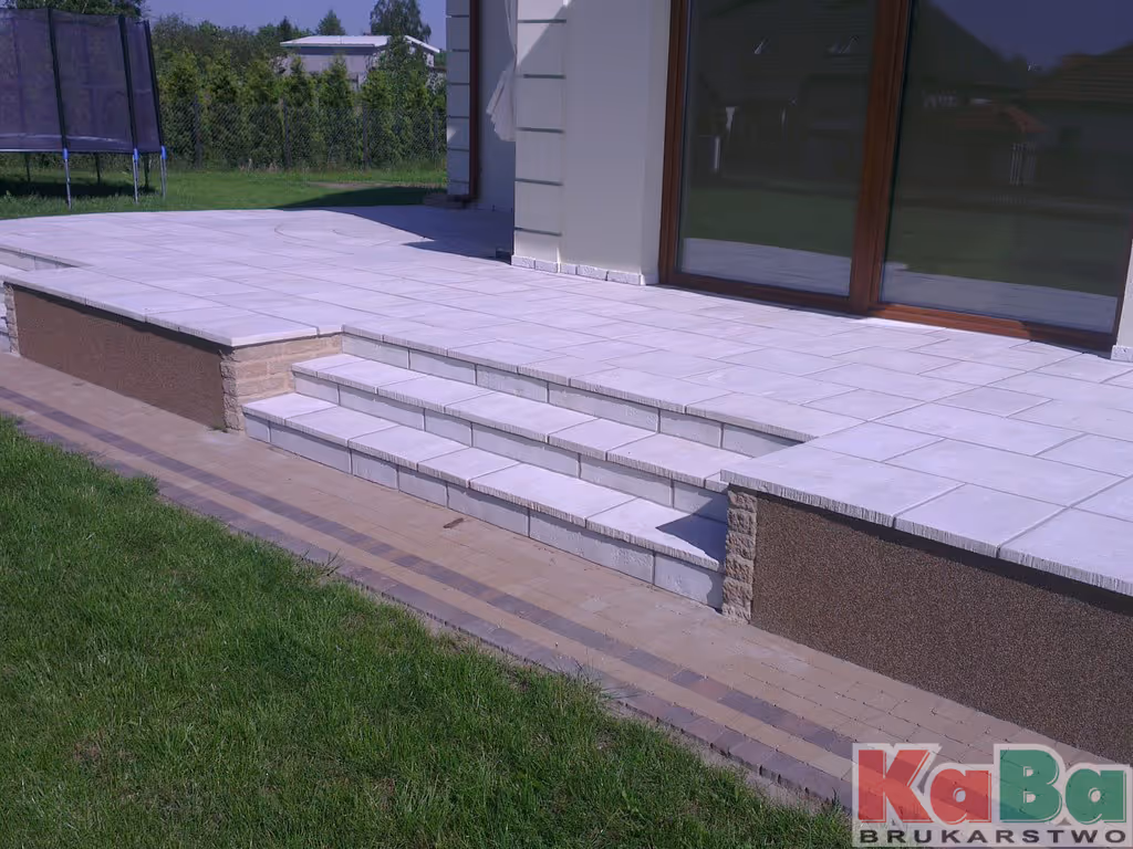 Taras z jasnych płytek z trzema stopniami prowadzącymi do ogrodu z zieloną trawą.