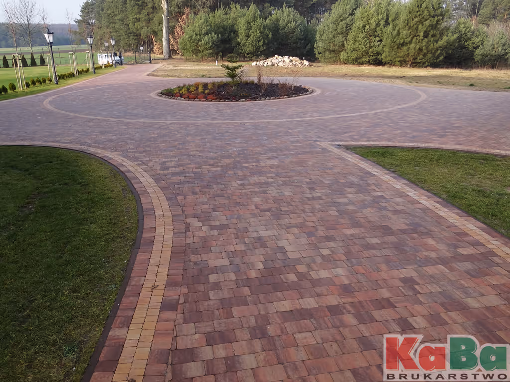 Brukowany chodnik z centralną okrągłą rabatą z roślinami, otoczony zielonymi trawnikami i drzewami w tle.