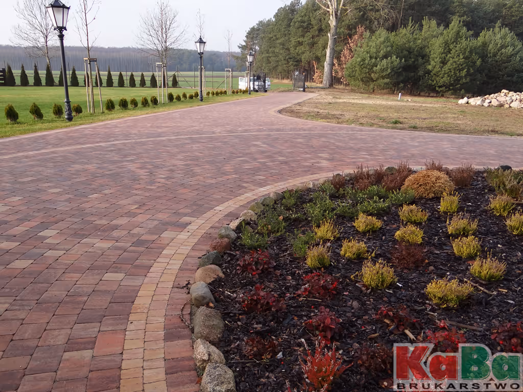 Wyłożona cegłą ścieżka biegnąca wzdłuż trawnika z latarniami i zaokrągloną rabatą z krzewami oraz drzewami w tle.