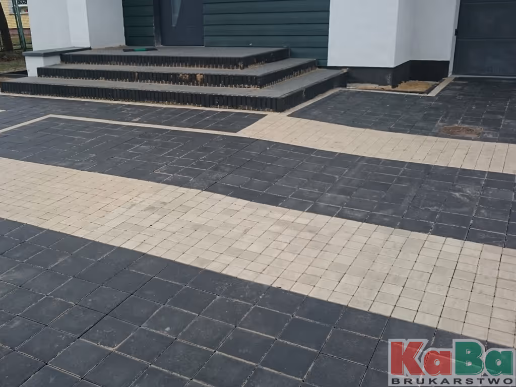 Schody i podjazd z ułożonymi czarno-białymi kostkami brukowymi przed wejściem do budynku.