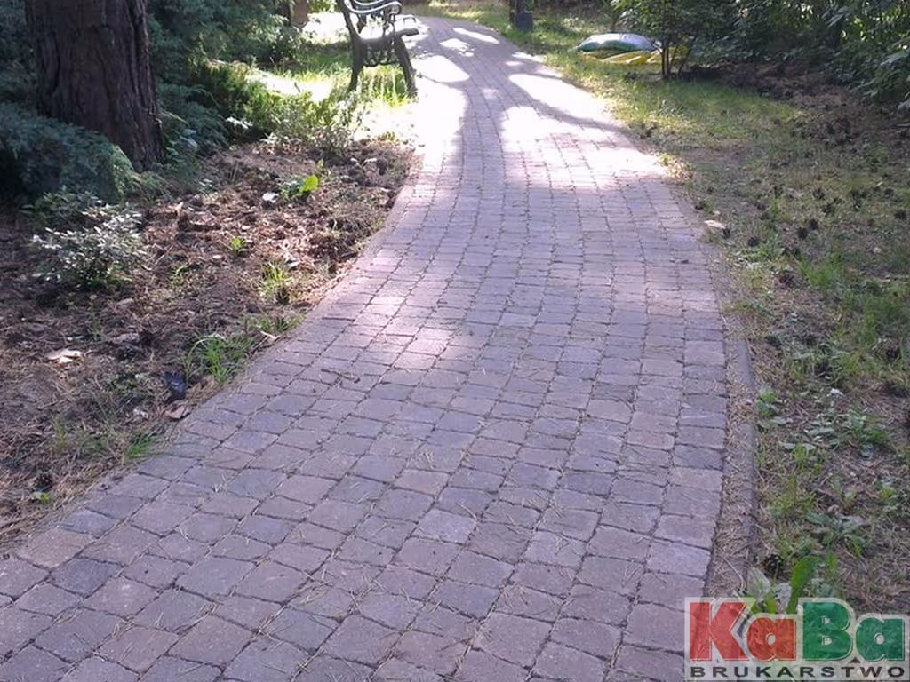 Kręta ścieżka z bruku w parku z ławką pod drzewami, częściowo oświetlona słońcem.