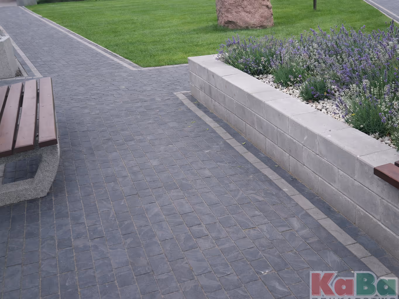 Ścieżka z ciemnoszarej brukowanej kostki z drewnianą ławką po lewej i podniesionym ogrodem z roślinami po prawej stronie.