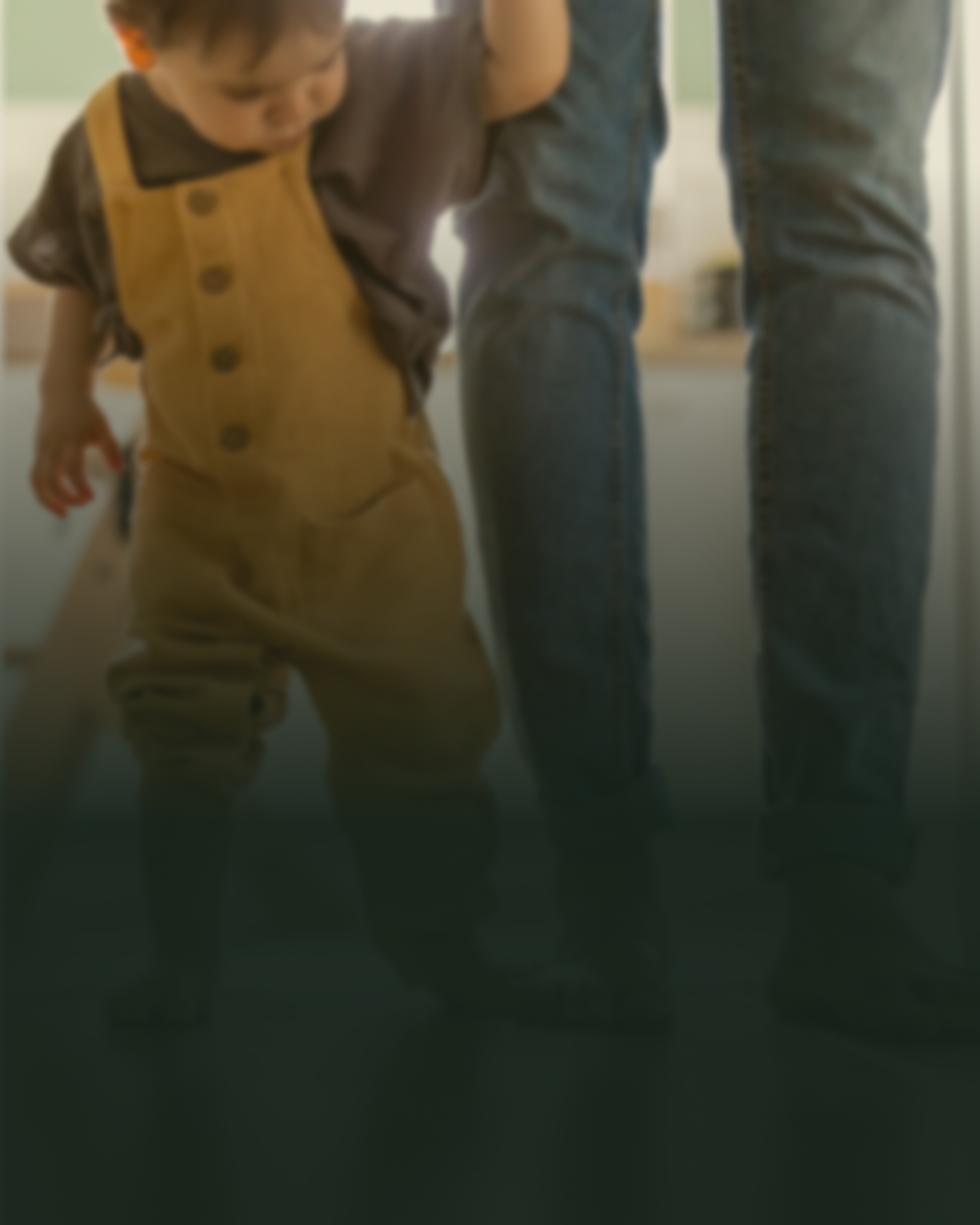 Lille barn går fremad indendørs med sin far i hånden. Der er en mørk skygge på billedet, som slører motivet en smule.