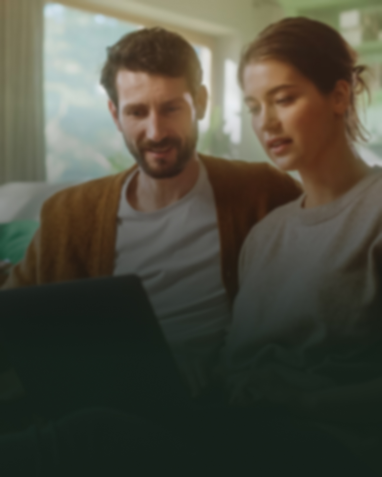 Mand og kvinde sidder og kigger på en computer i deres stue i hus. Der er en mørk skygge på billedet, som slører motivet en smule.