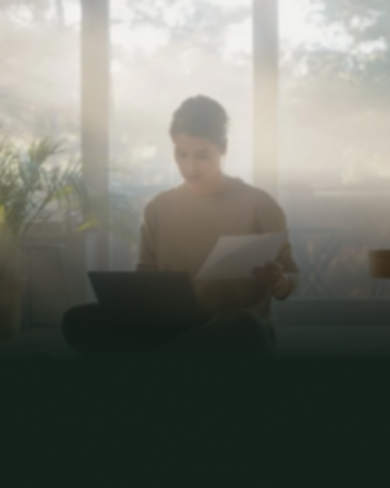 Kvinde sidder på gulvet med sin computer og holder et stykke papir. Der er en mørk skygge på billedet, som slører motivet en smule.