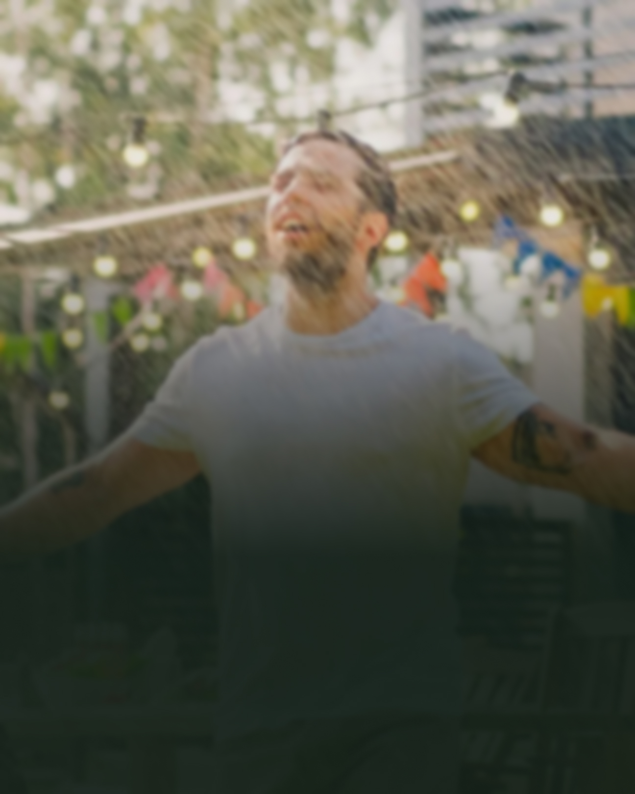 Mand smiler imens han bliver våd af regnvejr på en solrig dag. Der er en mørk skygge på billedet, som slører motivet en smule.